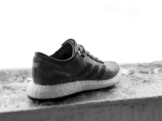 A single athletic shoe is placed on a ledge overlooking a blurred cityscape. The shoe is predominantly dark in color with a textured surface and a white, cushioned sole. The background is out of focus, emphasizing the shoe in the foreground.