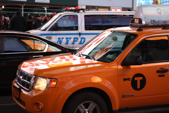 A yellow taxi is parked on the street, reflecting the bright lights of the surrounding urban environment. Nearby is an NYPD police vehicle, indicating a busy city setting.