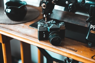 A vintage film camera sits on a wooden table next to an old sewing machine. The setting appears cozy with a muted color palette, suggesting a nostalgic or retro theme. A dark bowl is placed on a round mat nearby, highlighting a sense of simplicity in the composition.
