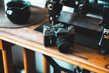 A vintage film camera sits on a wooden table next to an old sewing machine. The setting appears cozy with a muted color palette, suggesting a nostalgic or retro theme. A dark bowl is placed on a round mat nearby, highlighting a sense of simplicity in the composition.