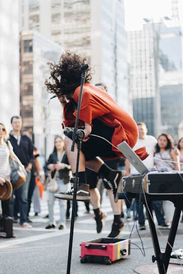 A performer with long curly hair is energetically jumping in the air while singing into a microphone. The scene captures a lively street performance with a small crowd gathered in the background. The performer is wearing a red top and black shorts. Various pieces of musical equipment, including a microphone stand and a keyboard, are visible.