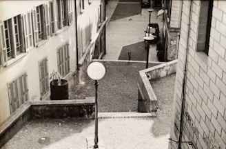 A black and white photograph of an urban street scene with cobblestone paving. The image captures a quiet, sloping alley with buildings on both sides. Windows with shutters and graffiti are visible on the walls. Street lamps line the path, and a single car is parked in the distance.