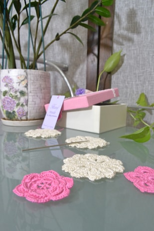 A glass table holds crocheted floral patterns in pink and off-white, with a metallic crochet hook resting on the table. To the side, there is a decorative ceramic pot with floral patterns containing a green leafy plant. A partially open pink box with a tag sits in the background, alongside a wall with a grayish texture.