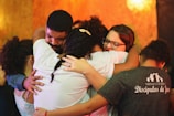 A group of people embracing each other closely in an emotional hug. Their expressions convey deep feelings, with some individuals having their eyes closed and holding tightly. The background is warm-toned, enhancing the heartfelt moment.