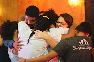 A group of people embracing each other closely in an emotional hug. Their expressions convey deep feelings, with some individuals having their eyes closed and holding tightly. The background is warm-toned, enhancing the heartfelt moment.