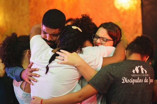 A group of people embracing each other closely in an emotional hug. Their expressions convey deep feelings, with some individuals having their eyes closed and holding tightly. The background is warm-toned, enhancing the heartfelt moment.