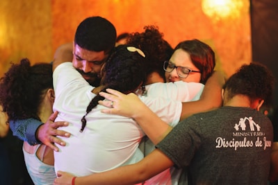 A group of people embracing each other closely in an emotional hug. Their expressions convey deep feelings, with some individuals having their eyes closed and holding tightly. The background is warm-toned, enhancing the heartfelt moment.