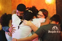A group of people embracing each other closely in an emotional hug. Their expressions convey deep feelings, with some individuals having their eyes closed and holding tightly. The background is warm-toned, enhancing the heartfelt moment.