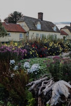 A picturesque garden with various colorful flowers in full bloom, including red, yellow, pink, and white blossoms. The garden is lush with green foliage and features some wooden logs in the foreground. Behind the garden, there is a charming house with a thatched roof and red-tiled sections. The sky is overcast, adding a serene and tranquil atmosphere.