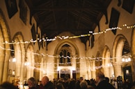 A warmly lit, historic church interior with high vaulted ceilings and stone arches. String lights are draped across the room, creating a festive atmosphere as people gather below, engaging in conversation. The ambiance suggests a community event or celebration, with a focus on the architectural beauty of the church.