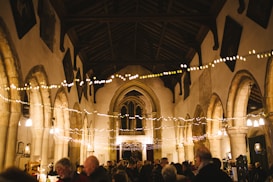 A warmly lit, historic church interior with high vaulted ceilings and stone arches. String lights are draped across the room, creating a festive atmosphere as people gather below, engaging in conversation. The ambiance suggests a community event or celebration, with a focus on the architectural beauty of the church.