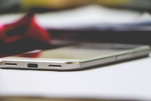 A smartphone lying on a flat surface, partially in focus, with its charging port and lower speaker grills visible. The background includes a blurred red and gray element, providing a contrast to the silver and black colors of the phone.