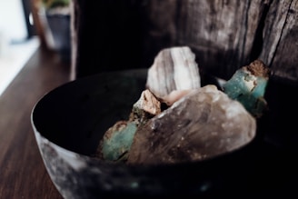 A close-up view of a collection of rough, unpolished stones in a dark, rustic bowl. The stones vary in color and texture, featuring shades of blue-green, brown, and clear crystal-like surfaces. The background is wooden, adding to the earthy and natural aesthetic of the scene.