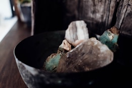 A close-up view of a collection of rough, unpolished stones in a dark, rustic bowl. The stones vary in color and texture, featuring shades of blue-green, brown, and clear crystal-like surfaces. The background is wooden, adding to the earthy and natural aesthetic of the scene.
