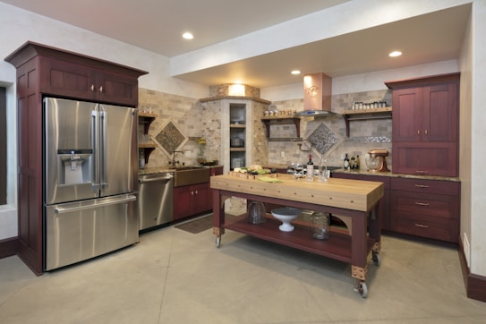 A modern kitchen features sleek stainless steel appliances, including a double-door refrigerator and a farmhouse sink. Dark wood cabinetry contrasts with the light-colored stone countertops and backsplash, creating a warm and inviting atmosphere. The center island is made of wood with shelves below and is topped with a cutting board. Various kitchen items and decorations, such as wine bottles, a mixer, and spice jars, are neatly arranged around the space.