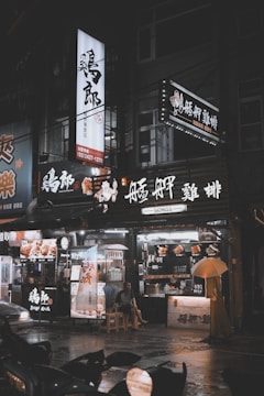 A dimly lit street scene featuring a storefront with illuminated signs in Chinese characters. The entrance has a person sitting on a stool under a large beige umbrella, surrounded by various signs and menus.