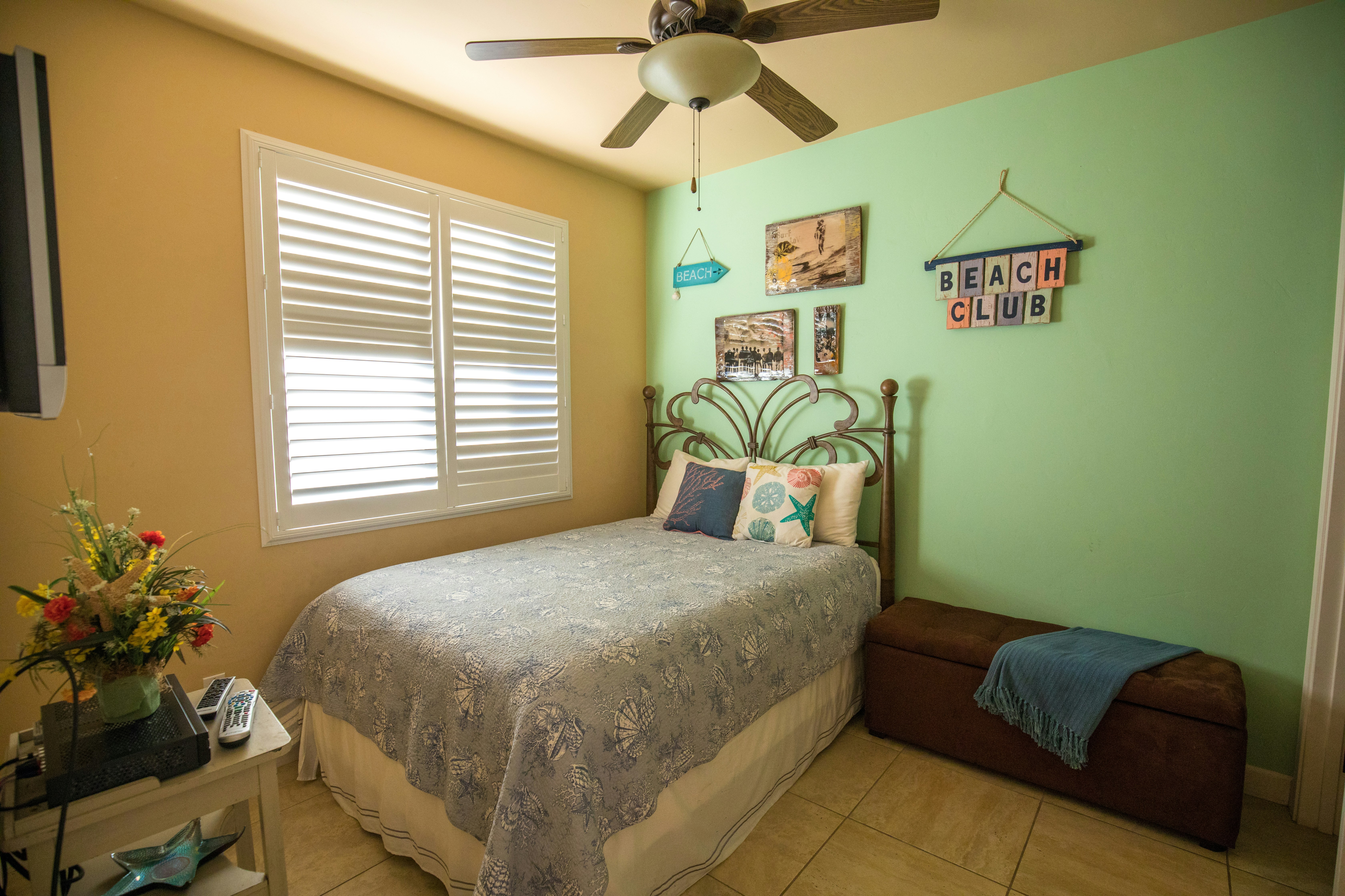 Bright beach-themed bedroom with iron bed, "Beach Club" wall sign, ceiling fan, and coastal décor.