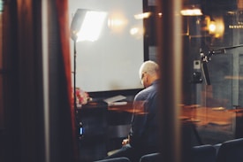 An older man sits alone in a dimly lit room, surrounded by professional lighting equipment and a microphone. The scene is viewed through glass, adding reflections that contribute to a sense of isolation. A table with some papers and a bouquet of flowers is in the background.