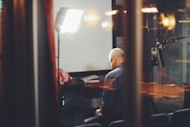 An older man sits alone in a dimly lit room, surrounded by professional lighting equipment and a microphone. The scene is viewed through glass, adding reflections that contribute to a sense of isolation. A table with some papers and a bouquet of flowers is in the background.