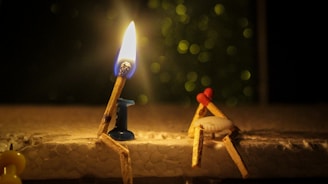 Two creatively arranged matchsticks are depicted sitting on a surface. One matchstick is lit with a bright flame, positioned in a small blue holder. The other matchstick appears to be leaning or seated against a white object, with a red element suggesting a head. The background is dark with blurred, circular light bokeh.