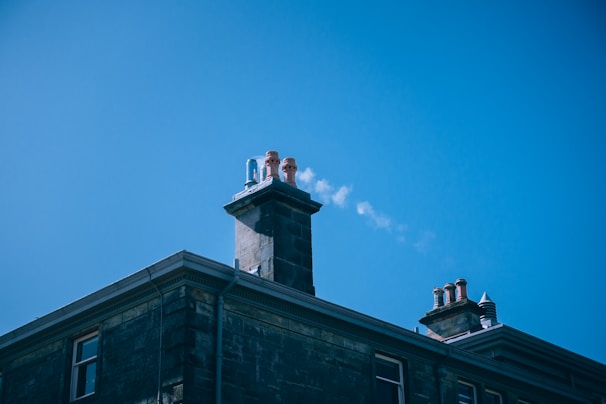A rooftop with a tall chimney releasing a small plume of smoke into the clear blue sky. The chimney is made of brick and there are multiple chimney pots on top. The building has a traditional architectural style with intricate stonework.