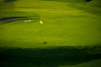 A green golf course with a yellow flag on a hole, surrounded by well-maintained grass. Shadows cast by the flag and surrounding landscape add contrast to the scene.