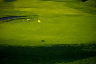 A green golf course with a yellow flag on a hole, surrounded by well-maintained grass. Shadows cast by the flag and surrounding landscape add contrast to the scene.