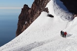 A group of mountaineers are trekking up a snowy slope on a mountain. The steep, rocky face of the mountain is visible to the left, contrasting with the bright white snow. The sky is visible in the background, showing a deep blue hue.