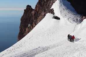 A group of mountaineers are trekking up a snowy slope on a mountain. The steep, rocky face of the mountain is visible to the left, contrasting with the bright white snow. The sky is visible in the background, showing a deep blue hue.