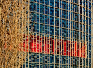 A high-rise building with a grid of blue-tinted glass windows reflecting the sky. Vertical beige columns run along the facade. In the background, a large red logo or sign is partially visible within the windows. A bare, leafless tree with intricate branches occupies the left side in the foreground.