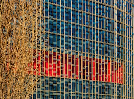 A high-rise building with a grid of blue-tinted glass windows reflecting the sky. Vertical beige columns run along the facade. In the background, a large red logo or sign is partially visible within the windows. A bare, leafless tree with intricate branches occupies the left side in the foreground.