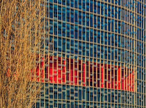 A high-rise building with a grid of blue-tinted glass windows reflecting the sky. Vertical beige columns run along the facade. In the background, a large red logo or sign is partially visible within the windows. A bare, leafless tree with intricate branches occupies the left side in the foreground.