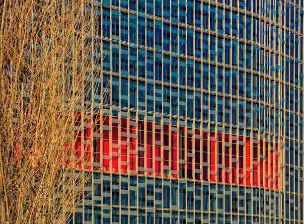 A high-rise building with a grid of blue-tinted glass windows reflecting the sky. Vertical beige columns run along the facade. In the background, a large red logo or sign is partially visible within the windows. A bare, leafless tree with intricate branches occupies the left side in the foreground.