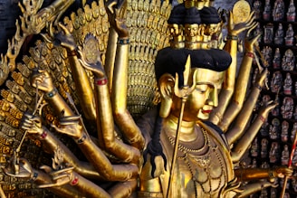 A golden statue featuring a multi-armed figure, likely a deity, adorned with intricate jewelry and details. The statue is set against a background filled with smaller, similarly styled figures. The craftsmanship showcases detailed facial features and ornate decorations on each arm.