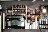 A café counter featuring a large espresso machine, stacks of white paper cups, bottles of flavored syrups, and a visible menu board. Shelves in the background hold coffee beans, wine bottles, and decorative items. The atmosphere appears to be cozy and inviting.