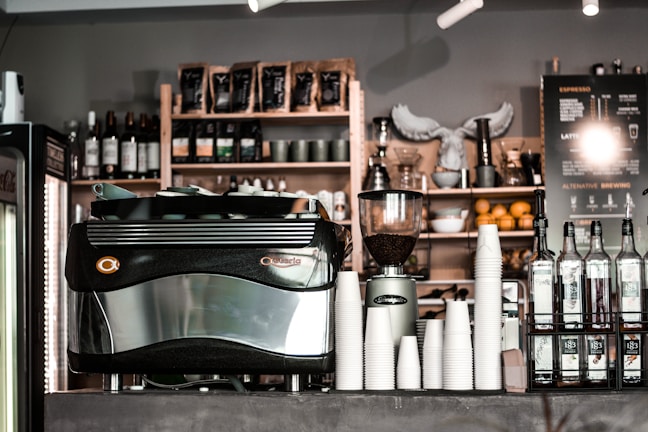 A café counter featuring a large espresso machine, stacks of white paper cups, bottles of flavored syrups, and a visible menu board. Shelves in the background hold coffee beans, wine bottles, and decorative items. The atmosphere appears to be cozy and inviting.