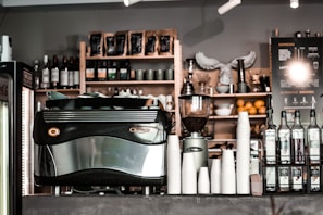 A café counter featuring a large espresso machine, stacks of white paper cups, bottles of flavored syrups, and a visible menu board. Shelves in the background hold coffee beans, wine bottles, and decorative items. The atmosphere appears to be cozy and inviting.