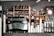 A café counter featuring a large espresso machine, stacks of white paper cups, bottles of flavored syrups, and a visible menu board. Shelves in the background hold coffee beans, wine bottles, and decorative items. The atmosphere appears to be cozy and inviting.