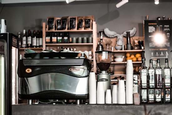 A caf&eacute; counter featuring a large espresso machine, stacks of white paper cups, bottles of flavored syrups, and a visible menu board. Shelves in the background hold coffee beans, wine bottles, and decorative items. The atmosphere appears to be cozy and inviting.