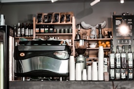 A café counter featuring a large espresso machine, stacks of white paper cups, bottles of flavored syrups, and a visible menu board. Shelves in the background hold coffee beans, wine bottles, and decorative items. The atmosphere appears to be cozy and inviting.