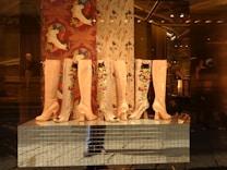 Several pairs of high-heeled boots with glossy finishes and colorful floral embroidery are displayed in a storefront. The background features decorative wallpaper with artistic bird designs. The reflection of a mannequin and interior lighting adds depth to the scene.