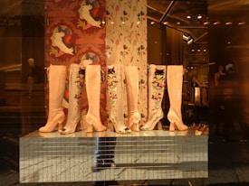 Several pairs of high-heeled boots with glossy finishes and colorful floral embroidery are displayed in a storefront. The background features decorative wallpaper with artistic bird designs. The reflection of a mannequin and interior lighting adds depth to the scene.