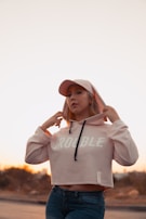 A person wearing a light pink hoodie with the word 'TROUBLE' printed on it, paired with blue jeans and a matching light pink cap. The person is adjusting the hoodie with both hands. The setting appears to be outdoors during a sunset, with a soft, warm glow in the background.