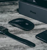 A collection of sleek, black electronic devices including a smartwatch, wireless mouse, smartphone, and a smartphone box on a wooden surface. The devices feature a minimalist design and the logo of a well-known technology brand.