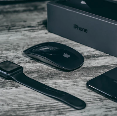A collection of sleek, black electronic devices including a smartwatch, wireless mouse, smartphone, and a smartphone box on a wooden surface. The devices feature a minimalist design and the logo of a well-known technology brand.