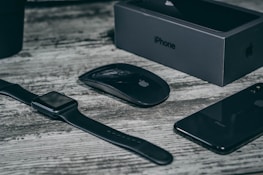 A collection of sleek, black electronic devices including a smartwatch, wireless mouse, smartphone, and a smartphone box on a wooden surface. The devices feature a minimalist design and the logo of a well-known technology brand.