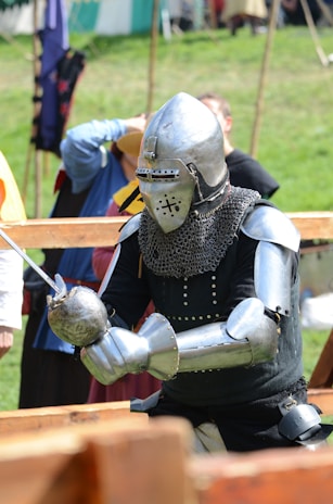 A person dressed in full medieval knight armor including a helmet and chainmail. They are holding a sword and standing outdoors in what appears to be a historical reenactment event. In the background, several other individuals in medieval or historical attire are visible.
