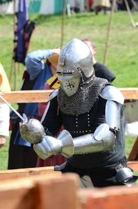 A person dressed in full medieval knight armor including a helmet and chainmail. They are holding a sword and standing outdoors in what appears to be a historical reenactment event. In the background, several other individuals in medieval or historical attire are visible.