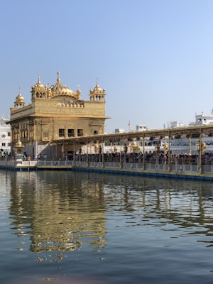 A stunning golden temple with intricate architecture is reflected in a serene body of water. The structure features multiple domes and ornate details. A long walkway leads to the temple, crowded with people. The sky is clear, enhancing the temple's grandeur.