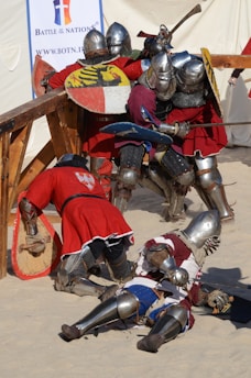 A group of armored knights engaged in a medieval-style battle in a sandy arena. They wear shiny metal helmets and chainmail, with bright red tunics over their armor. Some knights have fallen to the ground, while others clash with shields and swords. A banner in the background indicates an event named 'Battle of the Nations'.